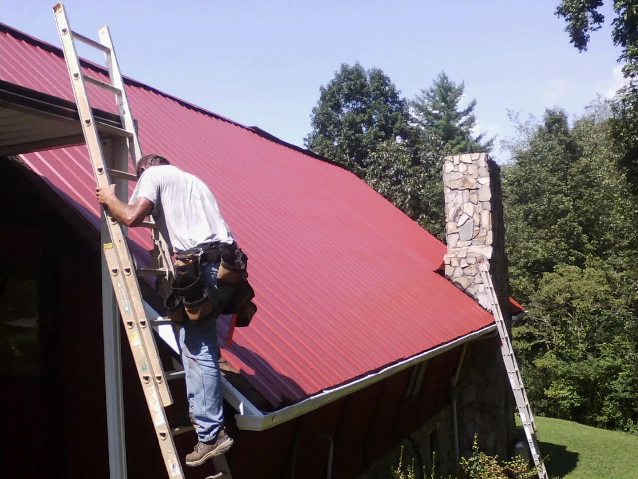Roofer on a ladder beside a red metal roof and stone chimney — Soffit & Fascia Repair by Roofing Brothers in Lemont